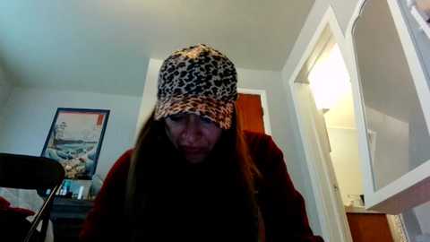 A video of a young woman with long, straight brown hair, wearing a leopard print bucket hat, standing in a dimly lit bedroom with white walls and a wooden door.