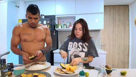 Video of a shirtless, muscular, tattooed Black man slicing avocado on a kitchen island, while a pale, brunette woman in a \"Boss\" T-shirt watches him, surrounded by avocado halves and other food items.