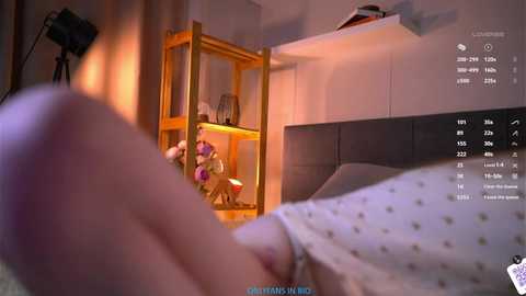 Video of a close-up hand holding a phone in a dimly lit bedroom, featuring a person wearing a white polka-dot nightgown, a wooden shelf with stuffed toys, and a digital clock showing 3:47 AM.