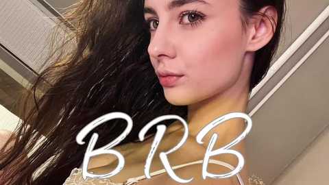 A close-up video of a young woman with long, wavy brown hair, light skin, and dark eyes. She has a soft, neutral expression. The background features a modern, beige-colored room with a ceiling light. Text \"B&R\" overlays her face.