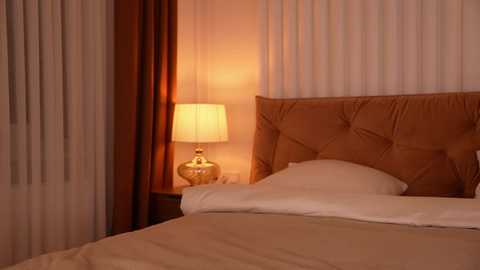 Video of a cozy, warmly lit bedroom featuring a tufted, brown headboard, beige bedspread, white pillows, a gold lamp with a white shade, and sheer curtains.