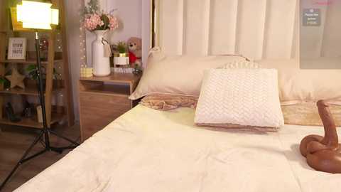 Media: Video of a cozy bedroom with cream-colored bedding, a plush pillow, and a wooden bear toy on the bed; background shows a wooden shelf, a potted plant, and a lamp.
