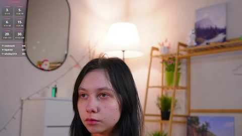 Video of an Asian woman with long black hair, wearing a nose ring, in a cozy room with a white lamp, a plant, and a calendar.