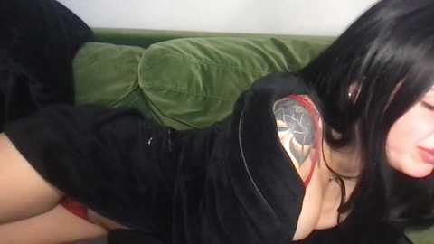 Video of a woman with long black hair, fair skin, and a black velvet dress with red lace details, lying on a green couch, partially visible.