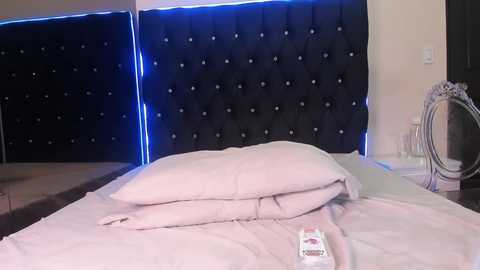 Video of a modern bedroom with a plush, tufted black headboard, a white bed with neatly folded pillows, a remote control on the bed, and a decorative mirror on the right wall.
