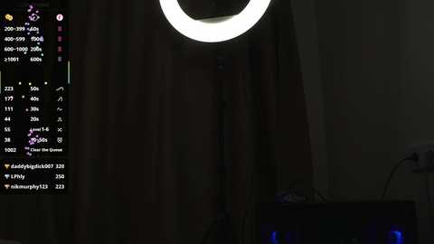 A dimly lit video of a room with a bright, circular light illuminating a man's face and upper torso. The background shows a curtain and a TV screen displaying a live stream of a gaming session.