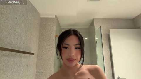 A video of an East Asian woman with straight black hair, fair skin, and light makeup, standing nude in a modern bathroom with beige tiles, glass door, and a towel bar.