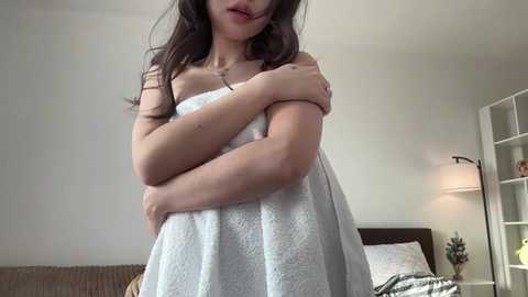 Video of a young woman with long dark hair, wearing a white towel, arms crossed, standing in a modern, minimalist bedroom with a bed and white wall-mounted shelf.