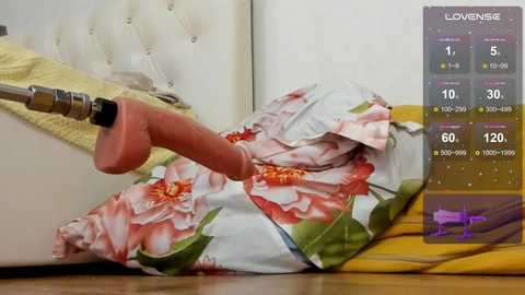 A video of a person lying on a white, tufted leather sofa, wearing a floral-patterned dress. A large, realistic pink dildo is penetrating them. The background includes a yellow rug and a digital screen displaying sex game statistics.