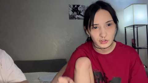 Video of a young woman with dark hair, wearing a red shirt, lying on a bed in a dimly lit room with a lamp and framed black-and-white photo on the wall.