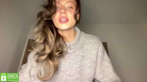 A video of a young woman with long, wavy blonde hair, wearing a light grey knitted sweater, standing indoors with a blurred background. She has a slightly open mouth, possibly mid-sentence.