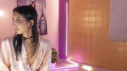 A video of a woman with long black hair and fair skin, wearing a silky pink robe, standing in a room with yellow and purple patterned walls.