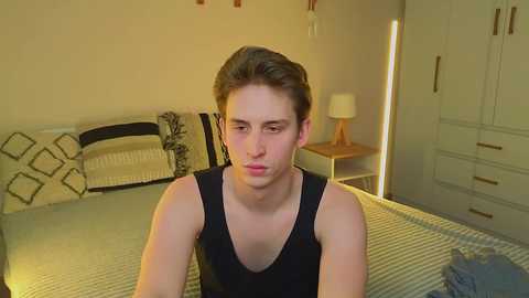 A video of a young, fair-skinned, androgynous person with short brown hair, wearing a black tank top, sitting on a neatly made bed with white and grey striped sheets and black geometric pillows in a beige room with a wooden nightstand and wardrobe.