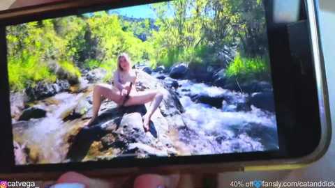 Video of a nude woman sitting on a rock in a forest stream, captured on a smartphone screen.