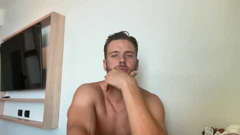 Video of a shirtless, light-skinned man with short brown hair, holding his chin thoughtfully against a plain white wall, with a wooden shelf and a flat-screen TV in the background.