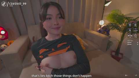 Video of a young woman with light skin, dark hair, and small breasts, lifting her shirt to reveal her nipples, in a dimly lit living room with a plush sofa, potted plants, and a red lamp.