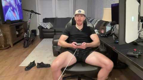 Video of a white man with long hair, wearing a beige cap and black shorts, playing a video game on a console, seated on a black chair in a cozy living room.