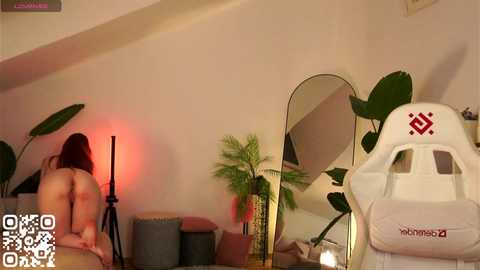 Video of a nude woman with fair skin, long brown hair, sitting on a gray couch, with a white gaming chair in the background.