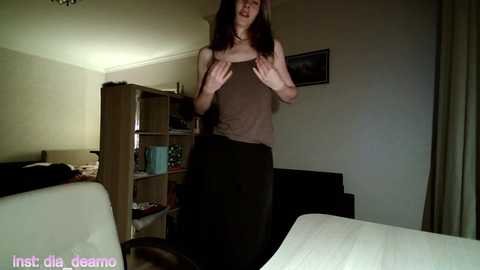 Media: A video of a woman in a dimly lit bedroom, wearing a brown tank top and black pants, standing in front of a wooden shelf with books and a blue mug.