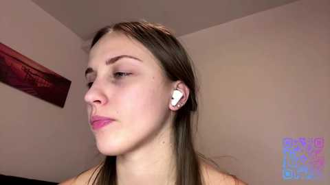 A close-up video of a young Caucasian woman with light skin, straight brown hair, and minimal makeup, wearing white earbuds. She has a neutral expression. The background features a beige wall and a red framed picture.