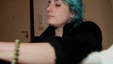 Video of a pale-skinned woman with teal hair, wearing a black robe, leaning against a beige wall, with a green bracelet on her wrist.