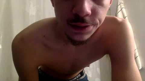 Video of a shirtless, light-skinned man with a mustache, slightly blurred, leaning forward with his lips parted, wearing blue jeans with a black waistband, in a dimly lit bathroom with white walls and a striped towel.