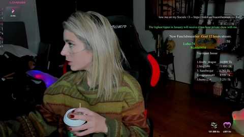 A video of a woman with straight, blonde hair in a green and brown striped sweater, holding a cup, seated in a dark room with a gaming chair and monitor displaying Twitch chat.