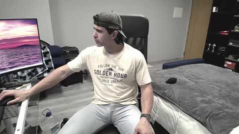 Media: Video of a young man in a casual beige T-shirt and light blue jeans, playing a video game on a monitor, in a modern, minimalist bedroom with a grey bed, a black chair, and a bookshelf.