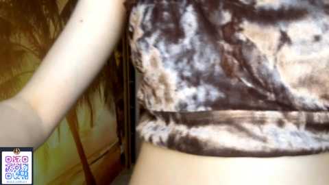 A video showing a close-up of a woman's bare midriff and side, wearing a tie-dye tank top, with her hand resting on a wooden surface. The background is blurred, emphasizing her attire.