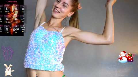 A video of a smiling, athletic, light-skinned woman with blonde hair, wearing a glittery, blue tank top and green shorts, dancing in a dimly lit room.