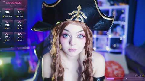 A video of a pale-skinned woman with long, wavy red hair, wearing a black pirate hat and makeup, set against a blurred background with a digital overlay of a game screen.