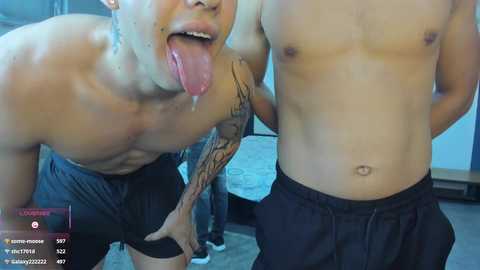 Video of two shirtless Asian men, one with a tattooed arm, licking each other's faces in a playful manner. They wear black shorts.