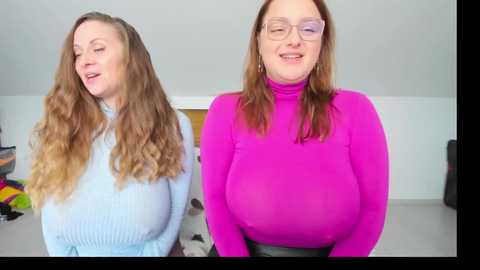 Video of two Caucasian women with long, wavy hair, one in a light blue sweater, the other in a bright pink turtleneck, standing in a brightly lit room with white walls and colorful clutter in the background.
