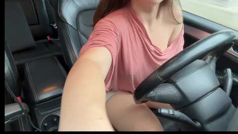 Video of a woman with long brown hair, wearing a pink off-shoulder top and white panties, sitting in a black leather car seat, holding the steering wheel.