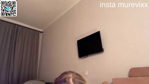 A video of a beige room with a flat-screen TV mounted on the wall, a person's head partially visible in the foreground, and a QR code in the top left corner.