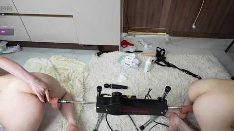 Video of a nude woman lying on a white carpet, being anally penetrated by a black sex machine. The room is cluttered with clothes and sex toys, including a dildo, on a wooden floor.