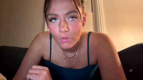 Media: Video of a young woman with light brown skin and dark hair, wearing a teal spaghetti-strap top, blowing a kiss. Background shows a dark couch and beige wall with a window.