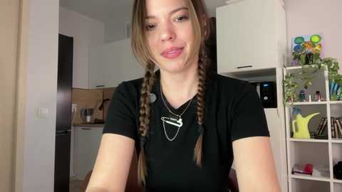 A video of a young woman with light skin, long brown hair in two braids, wearing a black t-shirt, smiling in a modern kitchen with white cabinets, a microwave, and potted plants.