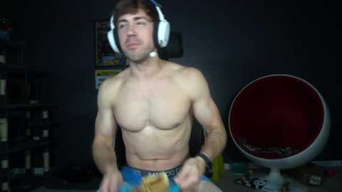 Video of a shirtless, muscular young man with light skin, brown hair, wearing headphones, playing a video game. Background includes dark walls, shelves, and a red beanbag chair.