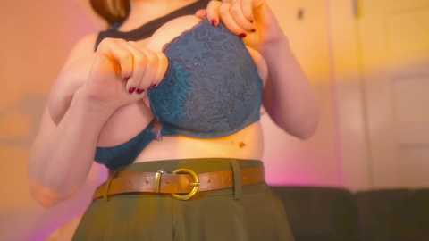 Video of a fair-skinned woman with brown hair, wearing a teal lace bra and olive-green skirt, adjusting her bra in a dimly lit room with warm hues.