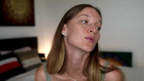 Video of a young Caucasian woman with light skin, brown hair, and freckles, wearing a green lace top, standing in a modern bedroom with minimalistic decor.
