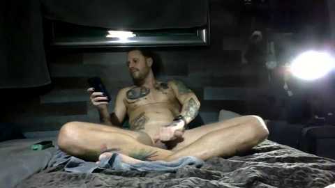 Media: Video of a tattooed, muscular, bearded man with long hair, sitting naked on a bed, holding a phone, in a dimly lit, rustic bedroom.