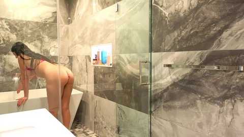 Media: Video of a woman with long black hair, wearing a pink bikini, bending over in a marble-tiled, glass-walled shower with a white bathtub.