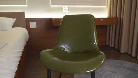 Video of a modern hotel room with a minimalist design. A green, leather, Eames-style chair is placed centrally. To the left, a neatly made bed with white linens and a wooden headboard is visible. A wooden desk with a beige curtain and a round rug add to the decor.