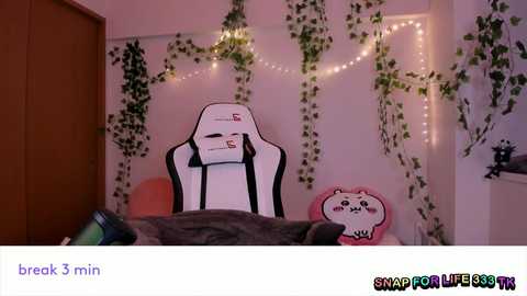 Video of a cozy, dimly-lit gaming room with a white gaming chair, a plush pink bear pillow, and fairy lights. The background is adorned with green ivy.