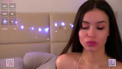 Video of a woman with long dark hair and fair skin, wearing a gold necklace, with closed eyes, resting on a beige cushioned headboard.