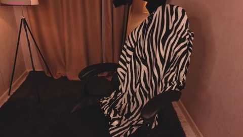Video of a person wearing a zebra-print shirt seated on a black sofa in a dimly lit room with beige walls, a tripod lamp, and a black trash can.