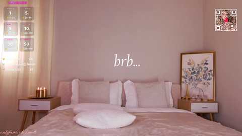 Video of a serene, minimalist bedroom with beige walls, a large bed, white pillows, fluffy pillows, and a floral painting. Soft, warm lighting, a wooden nightstand, and a small plant enhance the calming ambiance.