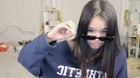Video of a young Asian woman with long black hair, wearing dark sunglasses and a blue sweatshirt with white text, adjusting her glasses in a cluttered, brightly lit bedroom with white walls and scattered toys.