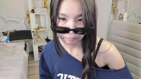 Video of an East Asian woman with long black hair, wearing a blue sweatshirt and black sunglasses, smiling in a cluttered bedroom with a white bed, yellow flowers, and a mirror.